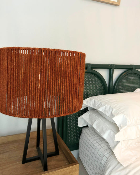 Handwoven lamp on a wooden nightstand next to a bed with white bedding.